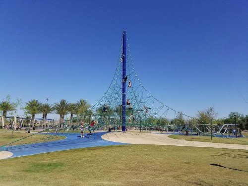 A large climbing structure in a park with children playing, surrounded by palm trees and a clear blue sky.
