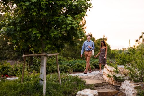 A couple walks hand in hand along a garden path, surrounded by greenery and trees during sunset.