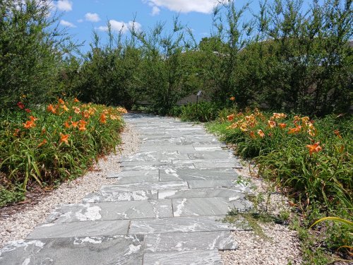 A stone pathway lined with vibrant orange flowers and greenery under a blue sky with scattered clouds.