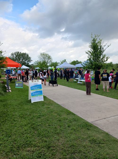 A community event with people walking along a path, surrounded by tents and greenery under a cloudy sky.