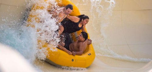 Three people in a yellow raft slide down a water slide, splashing water around them.