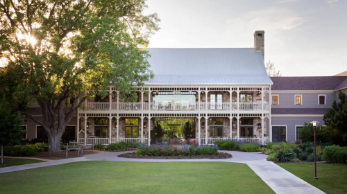 A large, elegant building with a wraparound porch, surrounded by greenery and a well-maintained lawn.