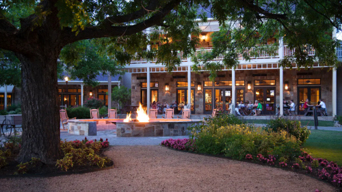 A cozy outdoor seating area with a fire pit, surrounded by lush greenery and blooming flowers, at dusk.