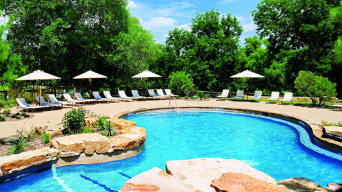 A serene pool area with lounge chairs and umbrellas surrounded by greenery under a bright blue sky.