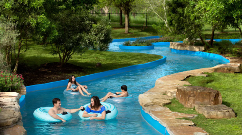 Four people float on inner tubes in a winding lazy river surrounded by greenery and stone pathways.