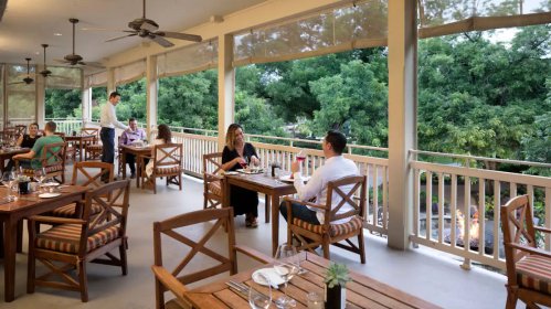 A restaurant patio with wooden tables, diners enjoying meals, and lush greenery in the background.