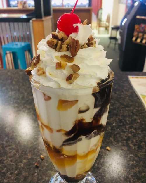 A tall glass dessert with layers of whipped cream, chocolate sauce, caramel, nuts, and a cherry on top.