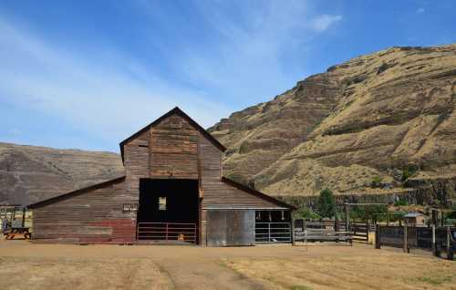 A rustic barn stands against a backdrop of dry, rolling hills under a blue sky. Fenced area in front.