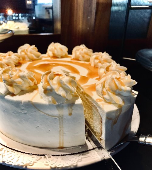 A creamy cake with caramel drizzle and decorative swirls, partially sliced on a decorative plate.