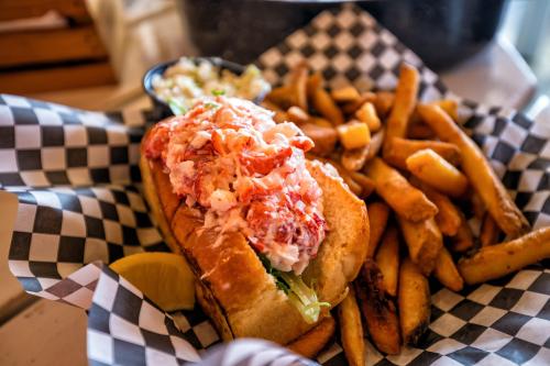 A lobster roll topped with lobster meat and lettuce, served with a side of fries and a lemon wedge on a checkered plate.