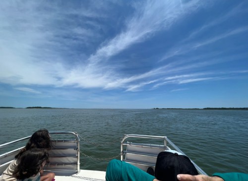 Two people sit on a boat, gazing at a calm waterway under a blue sky with wispy clouds.