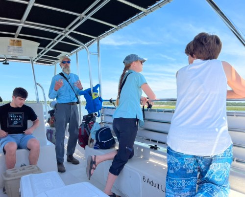 A group of people on a boat, with one man speaking while others look out at the water. Bright, sunny day.