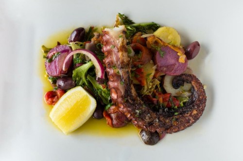 Grilled octopus served with vegetables, olives, and a lemon wedge on a white plate.