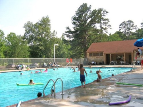 A sunny outdoor pool scene with children swimming, lounging on floats, and a wooden building in the background.