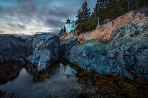 A lighthouse perched on rocky cliffs, reflecting in a calm tidal pool under a moody sky.