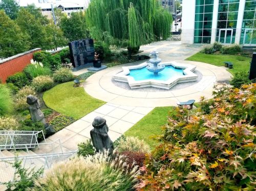 A serene garden with a central fountain, surrounded by sculptures, greenery, and a modern building in the background.