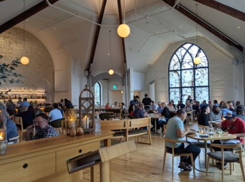 A bustling restaurant interior with high ceilings, wooden beams, and large windows, filled with diners at various tables.
