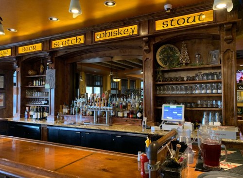 A wooden bar with shelves of liquor, taps, and a menu board displaying beer, scotch, whiskey, and stout.