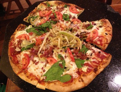 A pizza topped with tomatoes, lettuce, spinach, and bacon, with a few slices missing on a black table.
