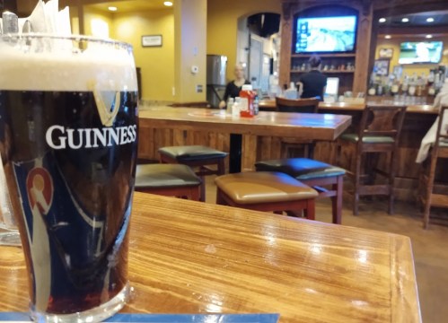 A pint of Guinness on a wooden table in a cozy bar with empty stools and a TV in the background.