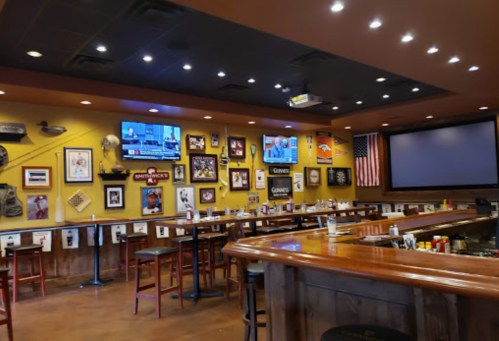 A cozy bar interior with a wooden counter, high stools, and walls adorned with sports memorabilia and a TV.