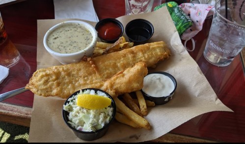 A plate with a large fried fish fillet, fries, coleslaw, and dipping sauces on a wooden table.