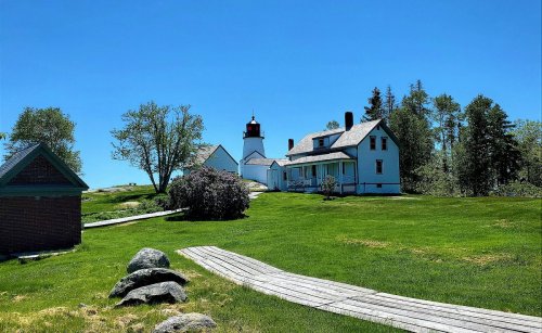 A scenic view of a lighthouse and a white house surrounded by green grass and trees under a clear blue sky.