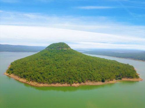 Aerial view of a lush, green island with a rounded hill, surrounded by calm blue water under a clear sky.