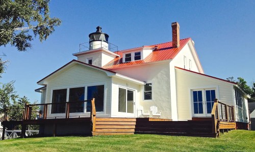 A white house with a red roof and lighthouse, featuring a deck and large windows, set against a clear blue sky.