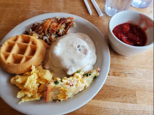 A plate of breakfast featuring a waffle, scrambled eggs, hash browns, sausage gravy, and a bowl of strawberry compote.