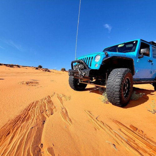 A blue Jeep Wrangler parked on a sandy desert landscape under a clear blue sky.