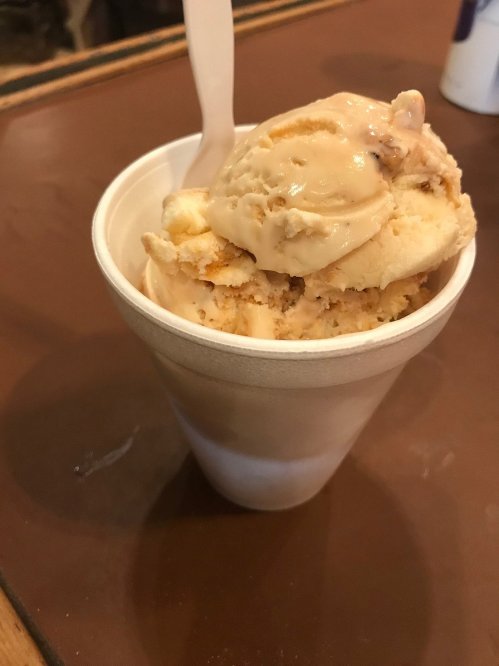 A cup filled with creamy ice cream, featuring a scoop of light brown and vanilla flavors, with a plastic spoon.