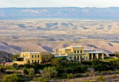 A yellow house sits on a hillside with a vast mountainous landscape in the background under a clear sky.