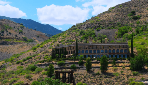 A large, historic building nestled on a hillside, surrounded by greenery and mountains under a blue sky.