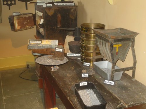 A wooden table displays various historical tools and materials, including a furnace, muller, and containers labeled for use.