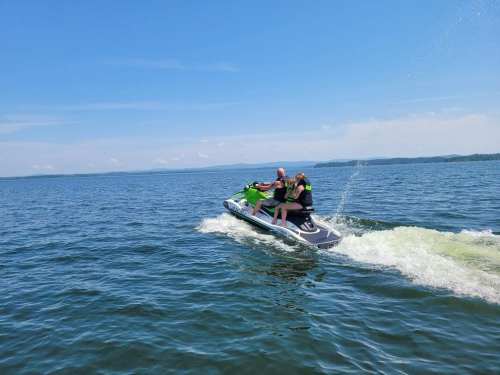 Two people ride a jet ski across a calm blue lake under a clear sky.