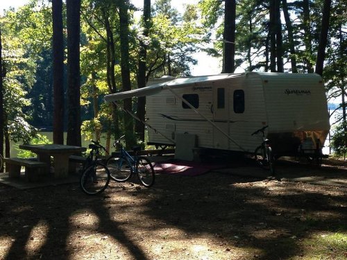 A camper trailer parked in a wooded area near a lake, with two bicycles and a picnic table nearby.