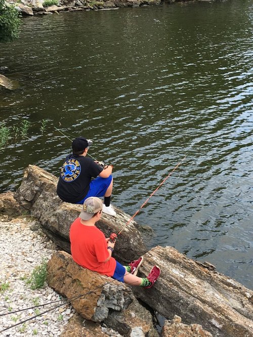 Two children sit on rocks by a river, fishing with rods, surrounded by greenery and calm water.