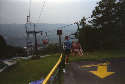 Two people sit on a ski lift, overlooking a scenic mountain view, with a path marked by an arrow in the foreground.