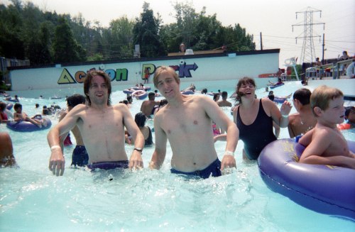 Two men wade in a water park pool, surrounded by people enjoying the water on a sunny day.