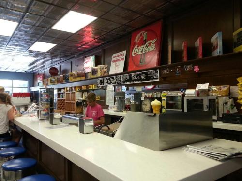 A retro diner interior with a white counter, blue stools, and vintage Coca-Cola signs, featuring a staff member serving.