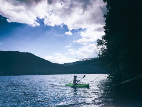 A person kayaking on a calm lake surrounded by mountains and trees under a partly cloudy sky.