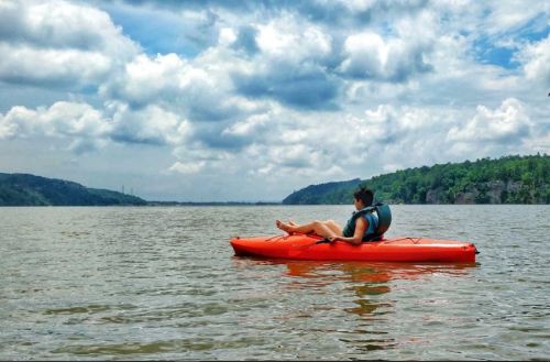 A person in an orange kayak paddles on a calm lake surrounded by green hills and a cloudy sky.