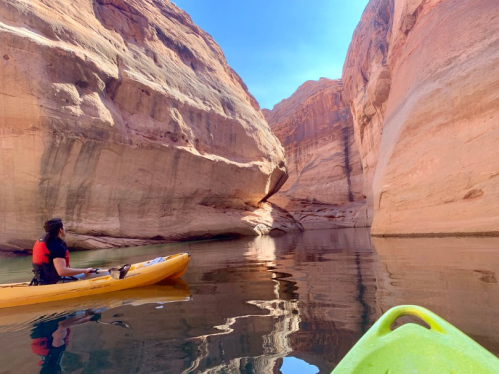 A person in a kayak paddles through a narrow canyon with towering rock walls and calm water reflecting the scenery.