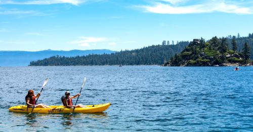 Two people kayak on a calm lake surrounded by trees and mountains under a clear blue sky.