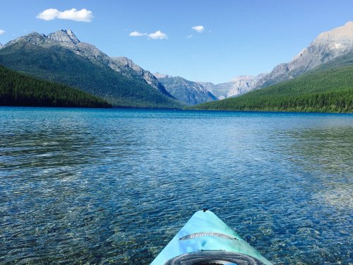A kayak on clear blue water, surrounded by mountains and lush green forests under a bright blue sky.