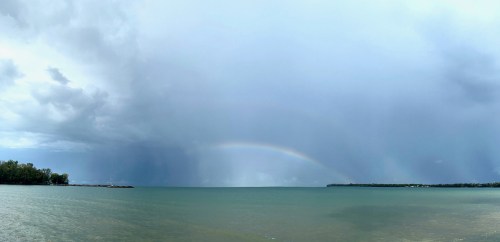 A panoramic view of a lake under a cloudy sky, featuring a faint rainbow arching over the water.