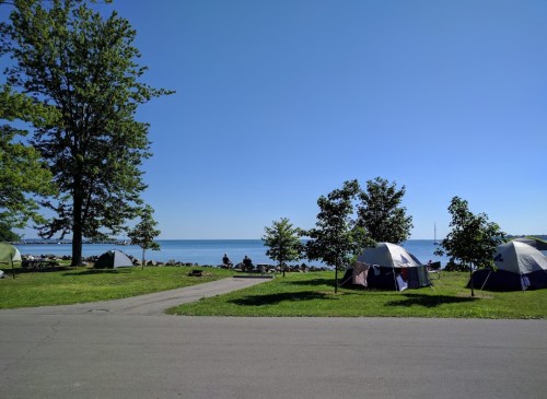 A serene lakeside view with tents set up on green grass, trees nearby, and a clear blue sky above.