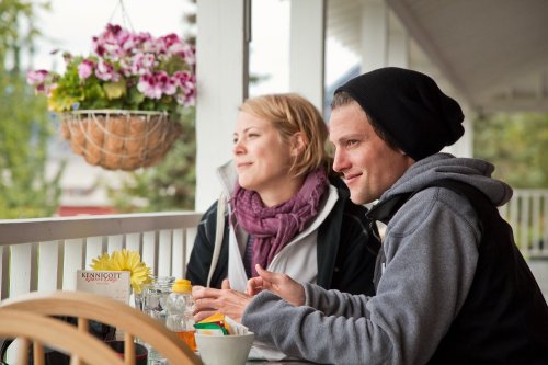 A man and woman sit at a table by a window, enjoying a meal and looking outside, with flowers in the background.
