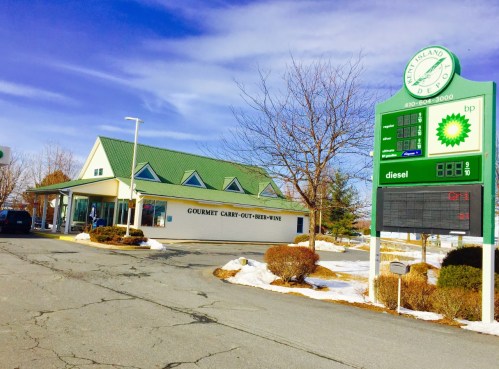 A convenience store with a green roof, featuring a sign for gourmet carry-out, beer, and wine, alongside a gas station.